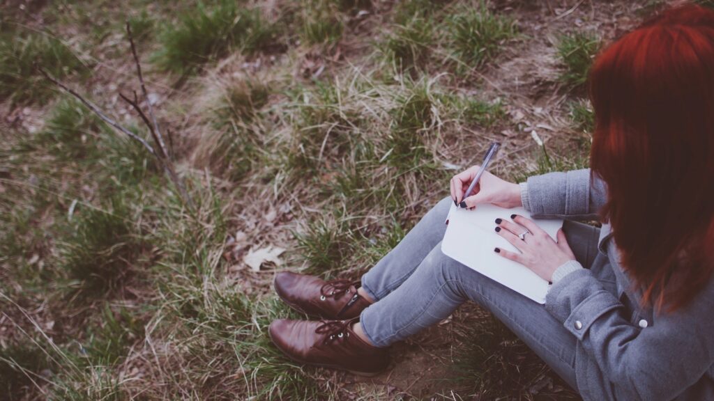 Redhead girl sitting on green spring grass writing down her thoughts in notebook.